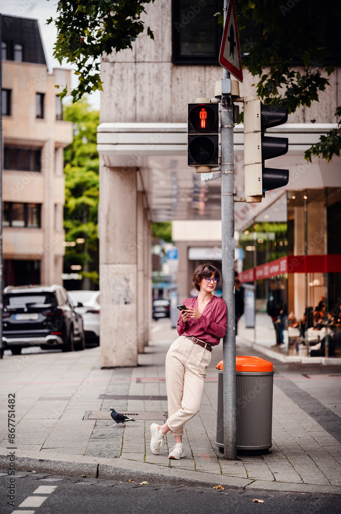 Woman in casual outfit leaning against a pole, checking her phone while waiting at a red pedestrian light in an urban area. Woman in casual outfit leaning against a pole, checking her phone while waiting at a red pedestrian light in an urban area.