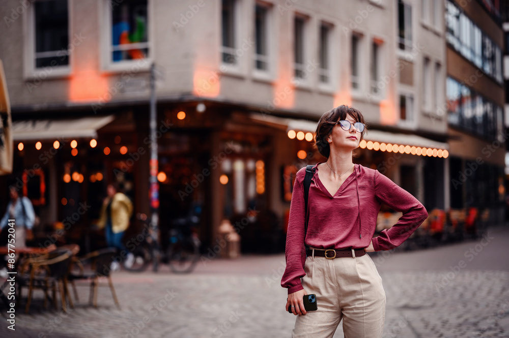 Fototapeta premium Woman in casual attire stands confidently with her hand on her hip, holding her phone, in a bustling city street with cafes and lights.