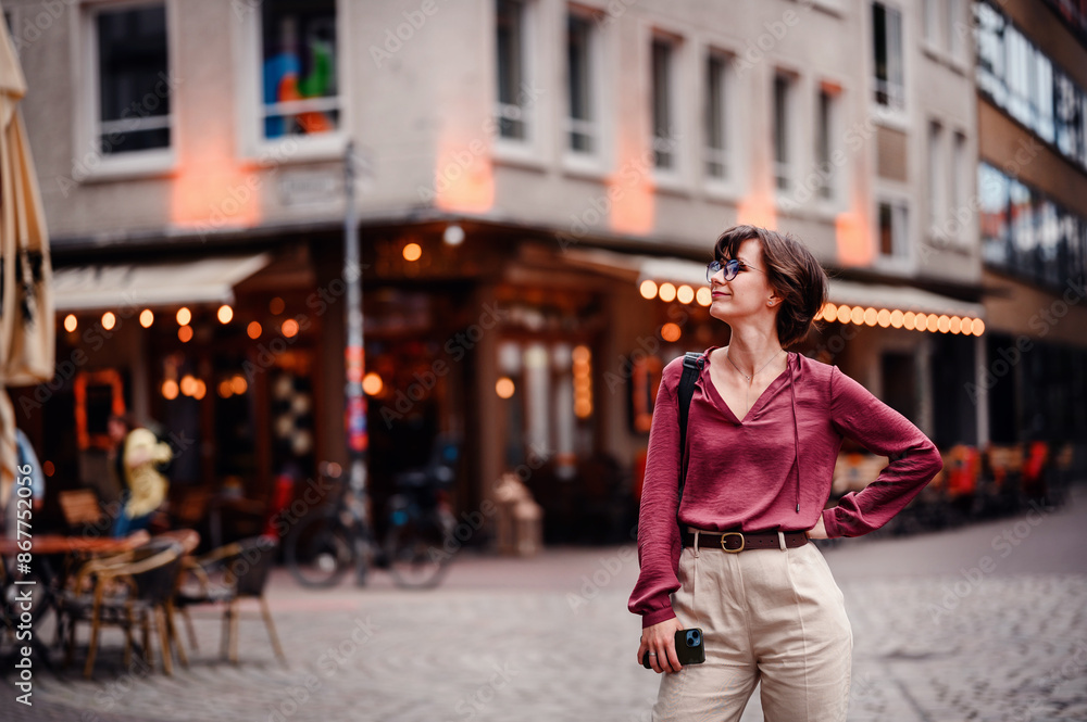 Fototapeta premium Woman in casual attire stands confidently with her hand on her hip, holding her phone, in a bustling city street with cafes and lights.