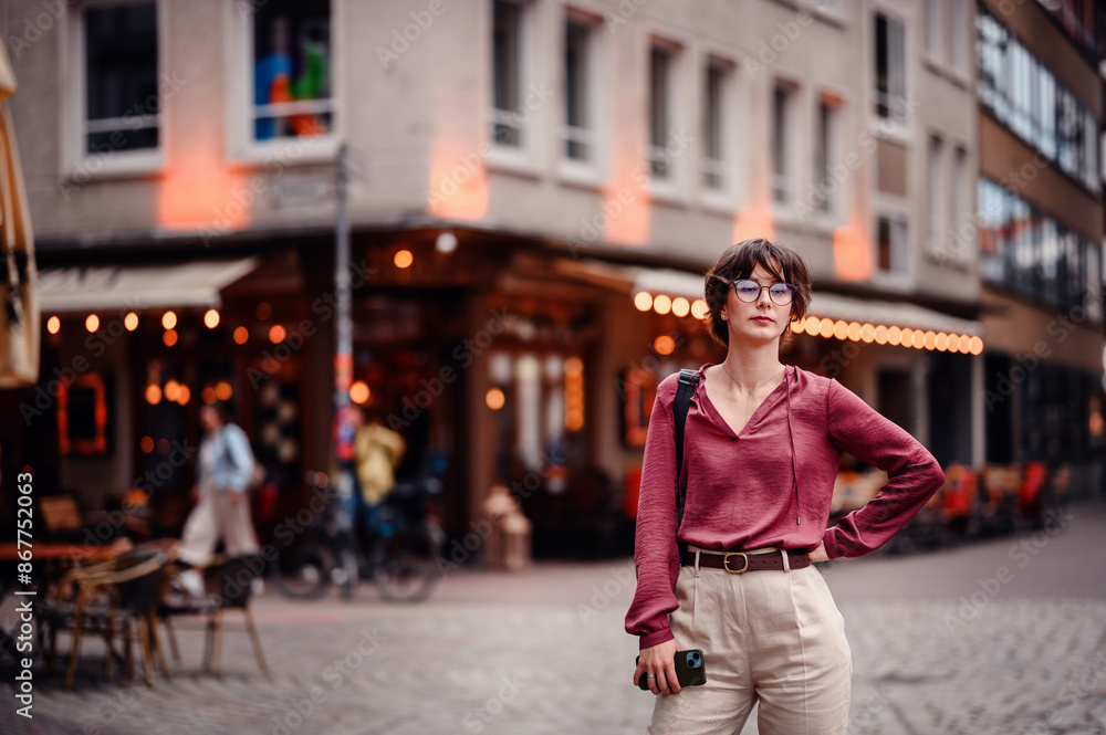 Fototapeta premium Woman in casual attire stands confidently with her hand on her hip, holding her phone, in a bustling city street with cafes and lights.