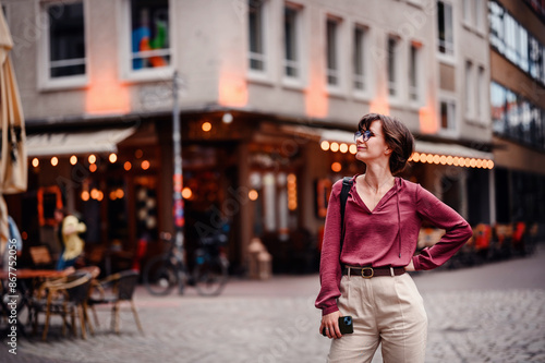 Woman in casual attire stands confidently with her hand on her hip, holding her phone, in a bustling city street with cafes and lights.