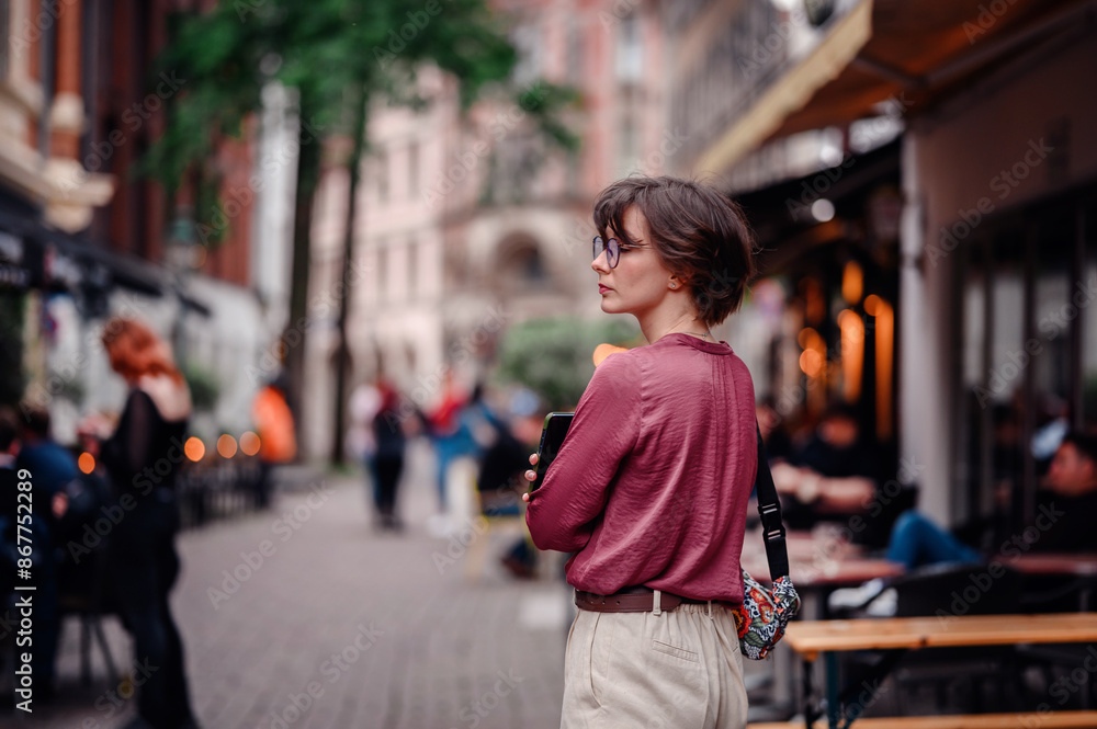 Woman in casual attire and glasses, holding her phone and backpack, exploring a bustling city street lined with cafes and people. Woman in casual attire and glasses, holding her phone and backpack, exploring a bustling city street lined with cafes and people.