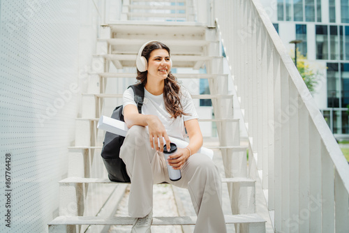 One young caucasian woman student sitting on stairs near university listening to music and drinking coffee