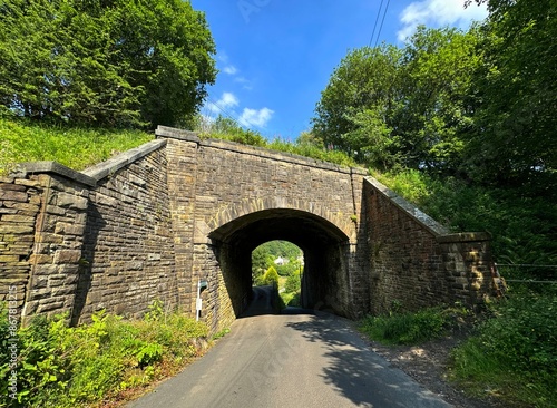 A rustic stone bridge arches over a narrow country road enveloped in lush greenery. Sunlight filters through the trees, casting dappled shadows over the road on, Brow Lane, Clayton, UK photo