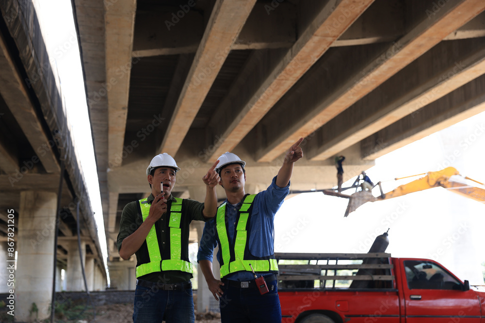 Two male construction engineers in safety vests and helmets discussing plans under the motorway bridge at the construction site Two male construction engineers in safety vests and helmets discussing plans under the motorway bridge at the construction site