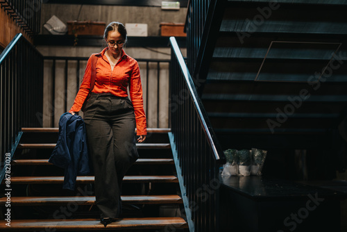 Young businesswoman in glasses and red blouse walking downstairs in a modern office building, holding a jacket. Contemporary workspace setting. © qunica.com