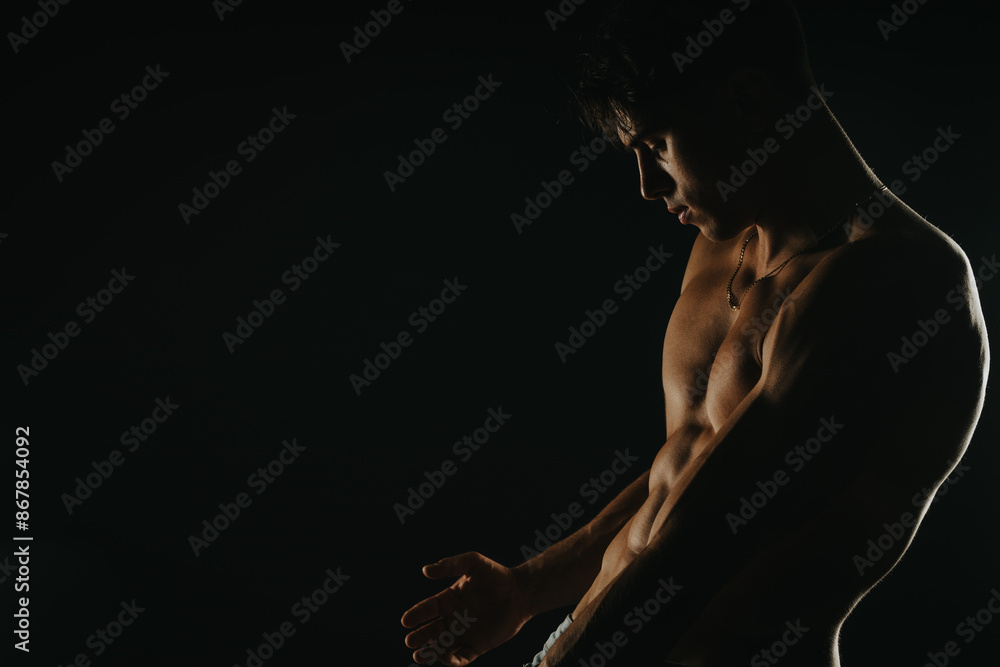 Shirtless muscular young man posing in low light, showcasing his defined physique and strength. Dark and dramatic lighting creates a moody and intense atmosphere. Shirtless muscular young man posing in low light, showcasing his defined physique and strength. Dark and dramatic lighting creates a moody and intense atmosphere.