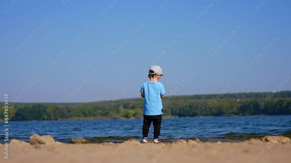 Rear view of a baby standing on the waterfront. Toddler kid throws the stones into water and picks new ones. Rear view of a baby standing on the waterfront. Toddler kid throws the stones into water and picks new ones.
