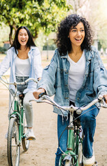 Vertical photo. Two young multiracial women having fun riding bike together in the park. Smiling female friends enjoying time together on summer vacation. Friendship and holidays concept.