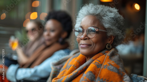 An elderly woman with grey hair and glasses smiles warmly as she sits outdoors with her friends, reflecting wisdom and contentment.
