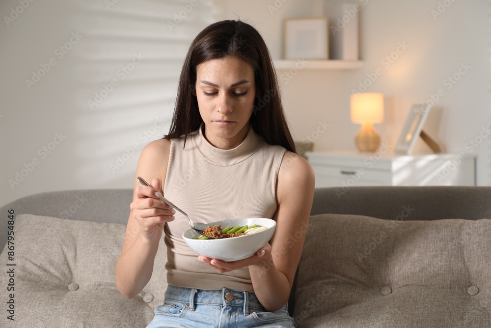 Eating disorder. Sad woman holding spoon with granola and bowl on sofa indoors Eating disorder. Sad woman holding spoon with granola and bowl on sofa indoors