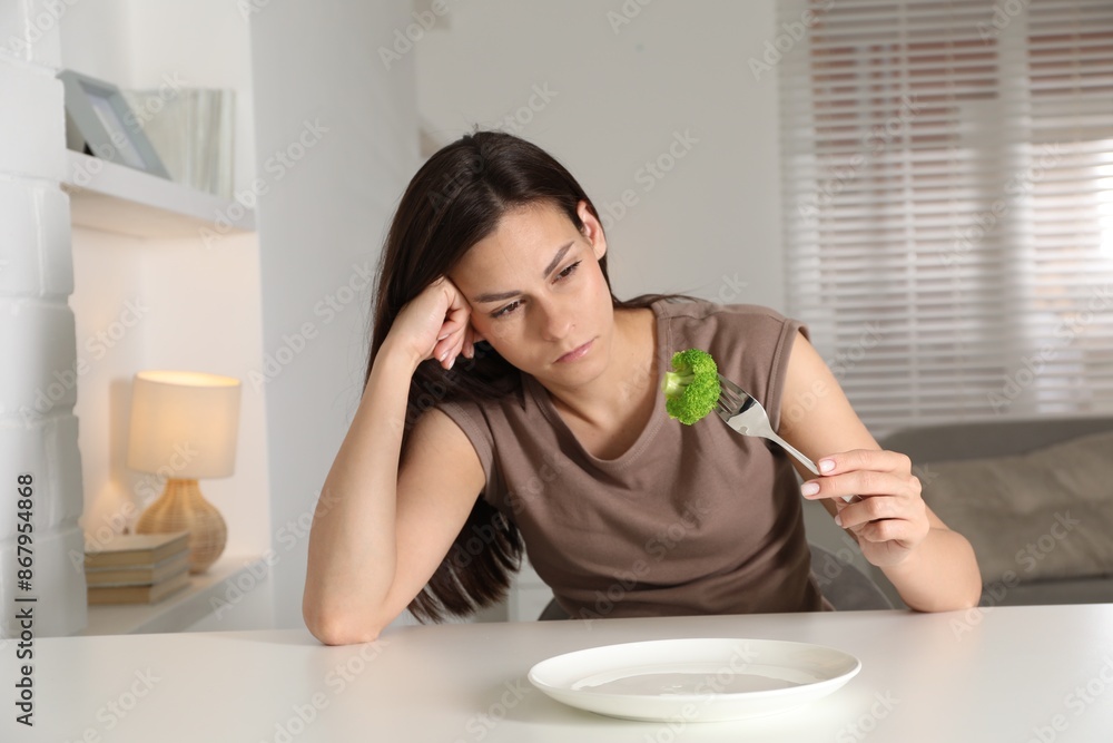 Eating disorder. Sad woman holding fork with broccoli at white table indoors Eating disorder. Sad woman holding fork with broccoli at white table indoors