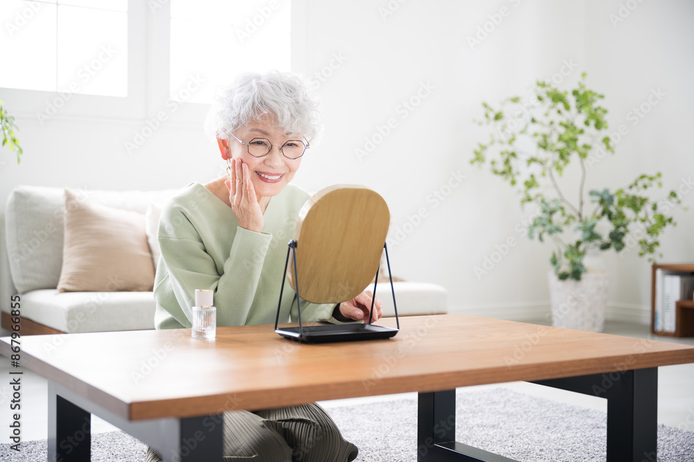 Senior (elderly) Japanese woman taking care of her skin (skin care) looking in the mirror in a brightly lit living room. Senior (elderly) Japanese woman taking care of her skin (skin care) looking in the mirror in a brightly lit living room.