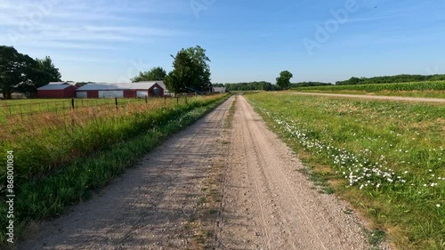 Slow motion views moving along a single lane dirt road in a rural area next to a farm. Agricultural buildings and corn fields surround the scene. Cloudy Blue skies overhead.
