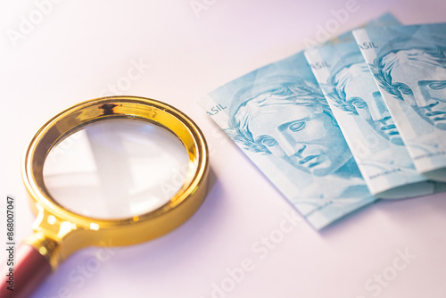 Dinheiro, Real - BRL, moeda do Brasil. 3 notas dobradas de 100 reais e uma lupa dourada. Fundo branco. Economia do Brasil, taxa de juros, inflação e desvalorização da moeda.