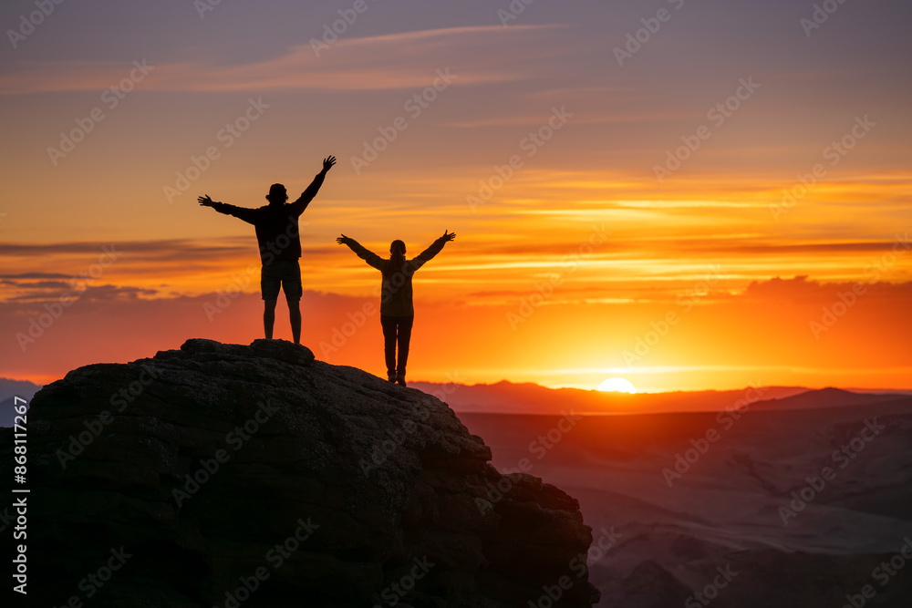 Two silhouettes of happy couple are standing with open arms on big rock against sunset sky and sun Two silhouettes of happy couple are standing with open arms on big rock against sunset sky and sun