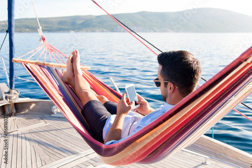 Photography Man with mobile phone relaxing on yacht in hammock