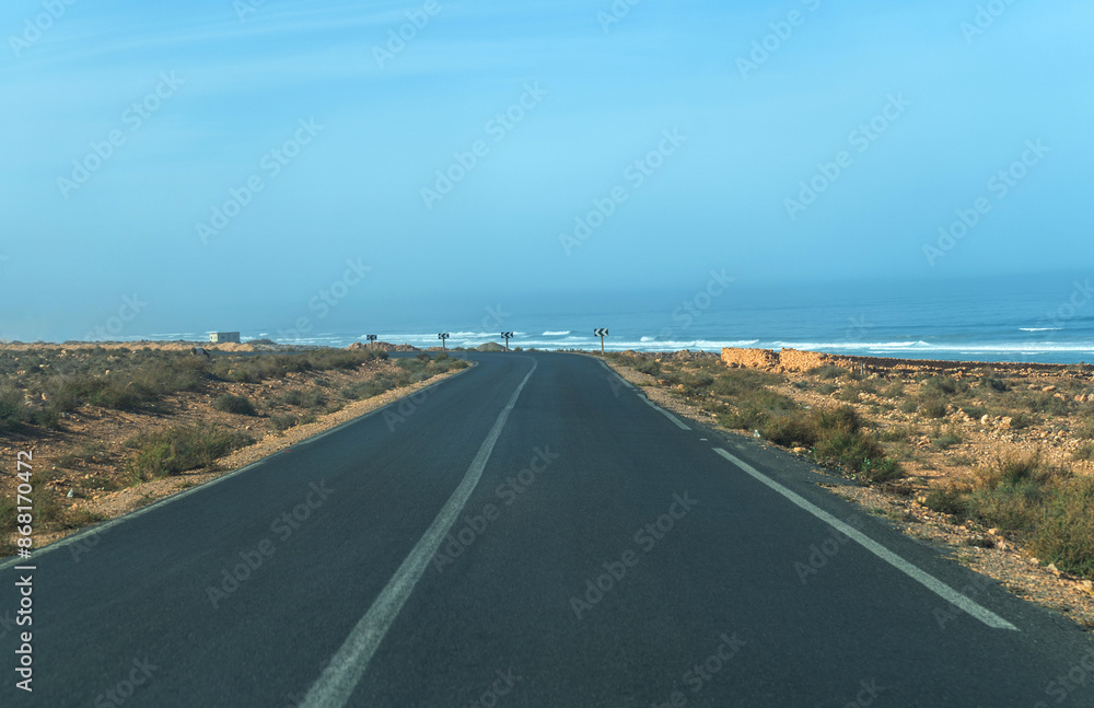 Fototapeta premium Road to ocean in Morocco, straight path, arid landscape, sparse vegetation, clear sky, distant waves