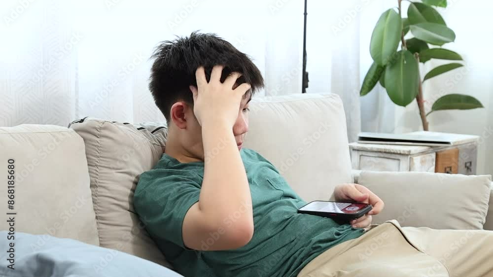 Young Asian teenage boy sitting on sofa rubbing eyes from eye strain. Smartphone on sofa next to him. Represents eye ache effects of excessive smartphone overuse among teenagers in a home environment Young Asian teenage boy sitting on sofa rubbing eyes from eye strain. Smartphone on sofa next to him. Represents eye ache effects of excessive smartphone overuse among teenagers in a home environment