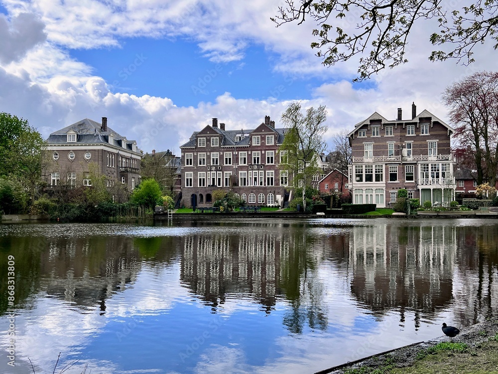 Naklejka premium Reflective Waters of Vondelpark: Historic Amsterdam houses reflect beautifully on the calm waters of Vondelpark, blending architecture with nature.