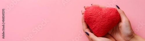 A hand holds a plush, red heart against a solid pink backdrop with copy space photo