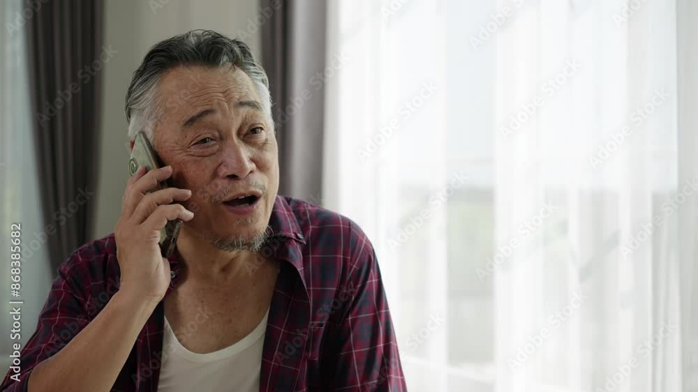 Handheld video of Smiling senior Asian man in plaid shirt talks on smartphone by bright window. Natural light illuminates his friendly expression. Casual home setting conveys warmth and connectivity. Handheld video of Smiling senior Asian man in plaid shirt talks on smartphone by bright window. Natural light illuminates his friendly expression. Casual home setting conveys warmth and connectivity.