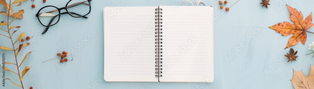 Open blank notebook with dried autumn leaves and eyeglasses on blue background. Ideal for seasonal writing, notes, and creative inspirations. Open blank notebook with dried autumn leaves and eyeglasses on blue background. Ideal for seasonal writing, notes, and creative inspirations.