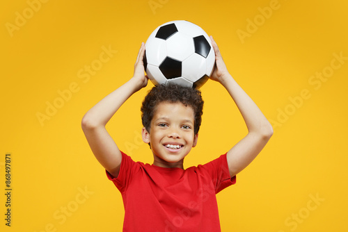 Studio portrait of happy smiling beautiful African American boy kid wearing basic red t-shirt, posing holding soccer ball over his head, isolated over bright colored orange yellow background