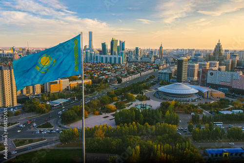 The Kazakhstan flag waiving above Astana city, with the panorama view of the city downtown, the Capital circus, green park, business buildings and skyscrapers, and residential buildings.
