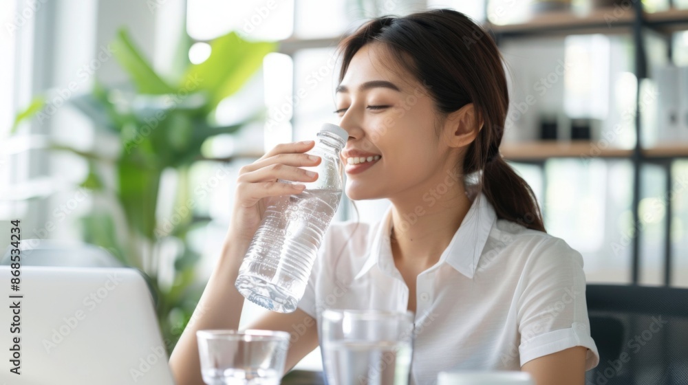 Smiling asian woman drinking water from a bottle while working at a desk with a laptop, surrounded by indoor plants. Smiling asian woman drinking water from a bottle while working at a desk with a laptop, surrounded by indoor plants.
