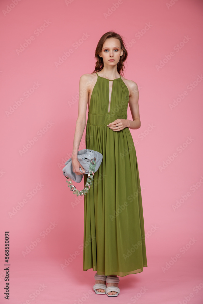 High fashion photo of a beautiful elegant young skinny woman in pretty long green dress, pastel blue handbag posing on pink background. Slim figure. High fashion photo of a beautiful elegant young skinny woman in pretty long green dress, pastel blue handbag posing on pink background. Slim figure.