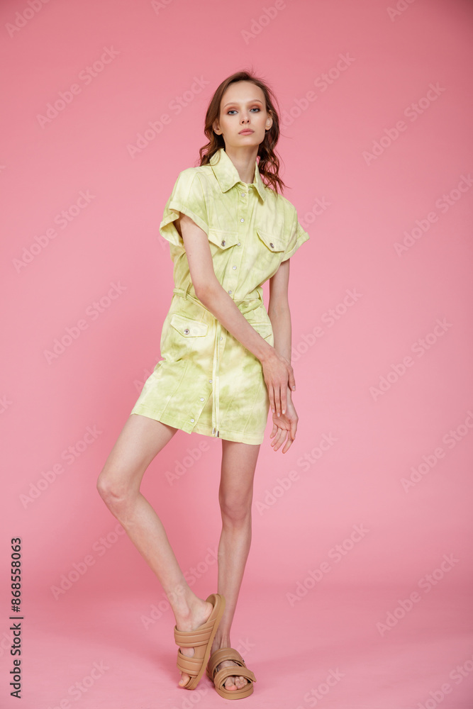High fashion photo of a beautiful skinny young woman in pretty green military denim jeans dress posing on pink background. Slim figure. High fashion photo of a beautiful skinny young woman in pretty green military denim jeans dress posing on pink background. Slim figure.