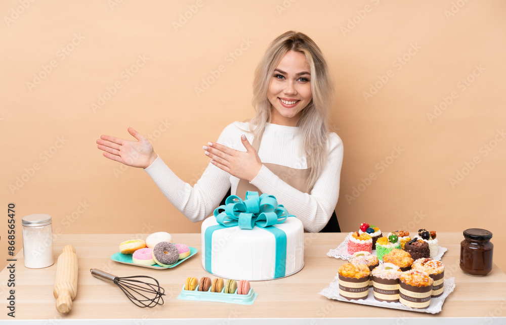 Teenager pastry chef with a big cake in a table extending hands to the side for inviting to come Teenager pastry chef with a big cake in a table extending hands to the side for inviting to come