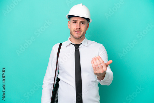 Young architect caucasian man with helmet and holding blueprints isolated on blue background inviting to come with hand. Happy that you came