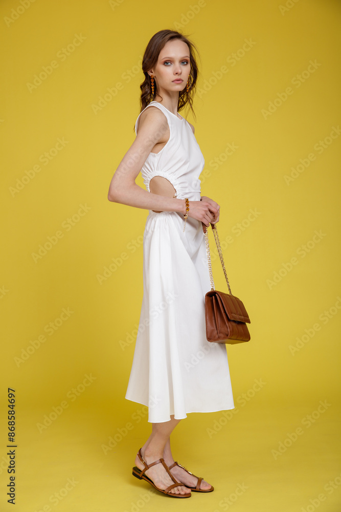 High fashion photo of a beautiful skinny young woman in pretty white dress, brown handbag posing on yellow background. Slim figure. High fashion photo of a beautiful skinny young woman in pretty white dress, brown handbag posing on yellow background. Slim figure.