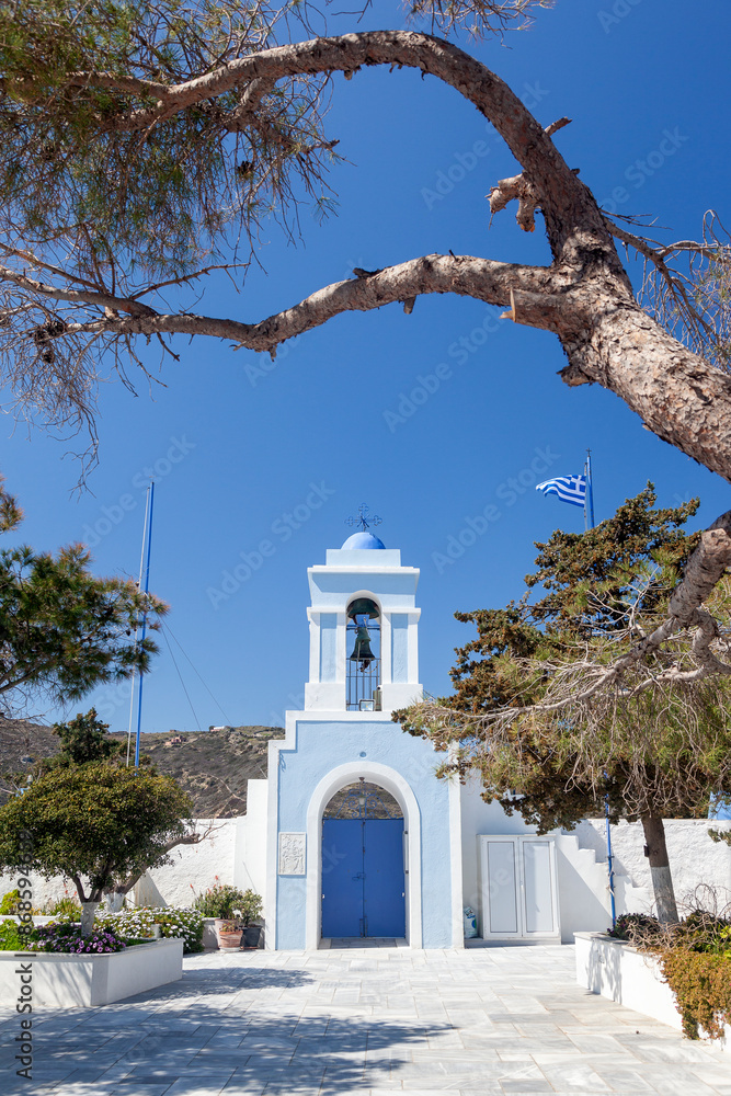 The beautiful Greek chapel of Panagia Kastriani, in Kea island, with a blue dome and bell tower, framed by trees against a clear blue sky, showcasing traditional Cycladic architecture.