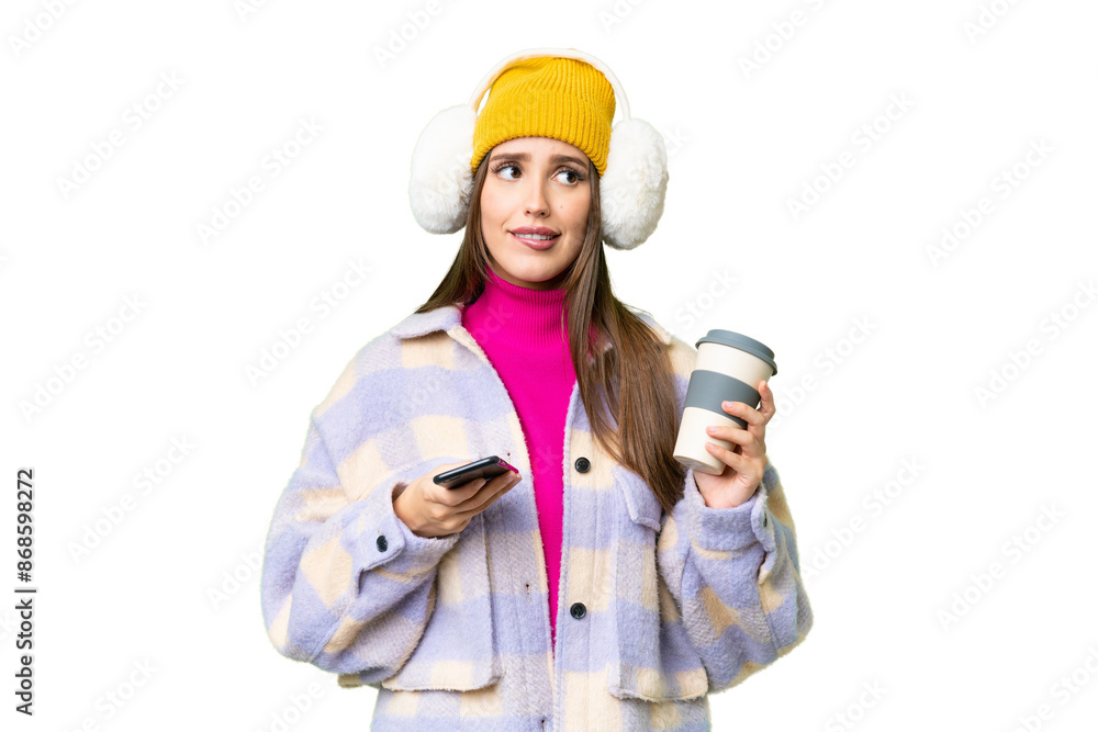 Young woman wearing winter muffs over isolated chroma key background holding coffee to take away and a mobile while thinking something Young woman wearing winter muffs over isolated chroma key background holding coffee to take away and a mobile while thinking something