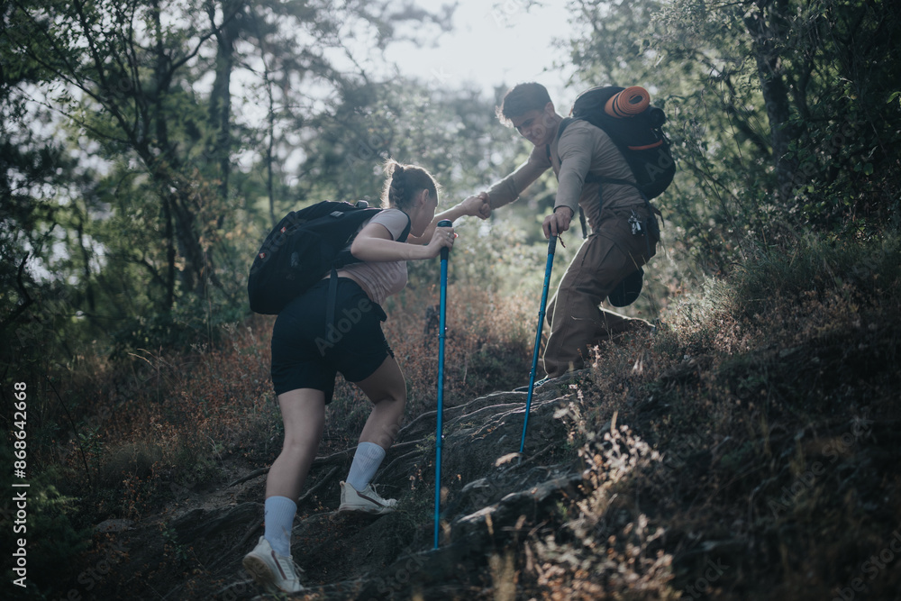 Two hikers helping each other ascend a steep trail in a forest, showcasing teamwork and outdoor adventure. Two hikers helping each other ascend a steep trail in a forest, showcasing teamwork and outdoor adventure.