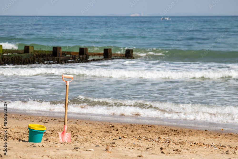 An image of colorful beach toys including a bucket and spade planted in the golden sandy shore, with gentle waves approaching and a clear, calm sea in the backdrop. An image of colorful beach toys including a bucket and spade planted in the golden sandy shore, with gentle waves approaching and a clear, calm sea in the backdrop.
