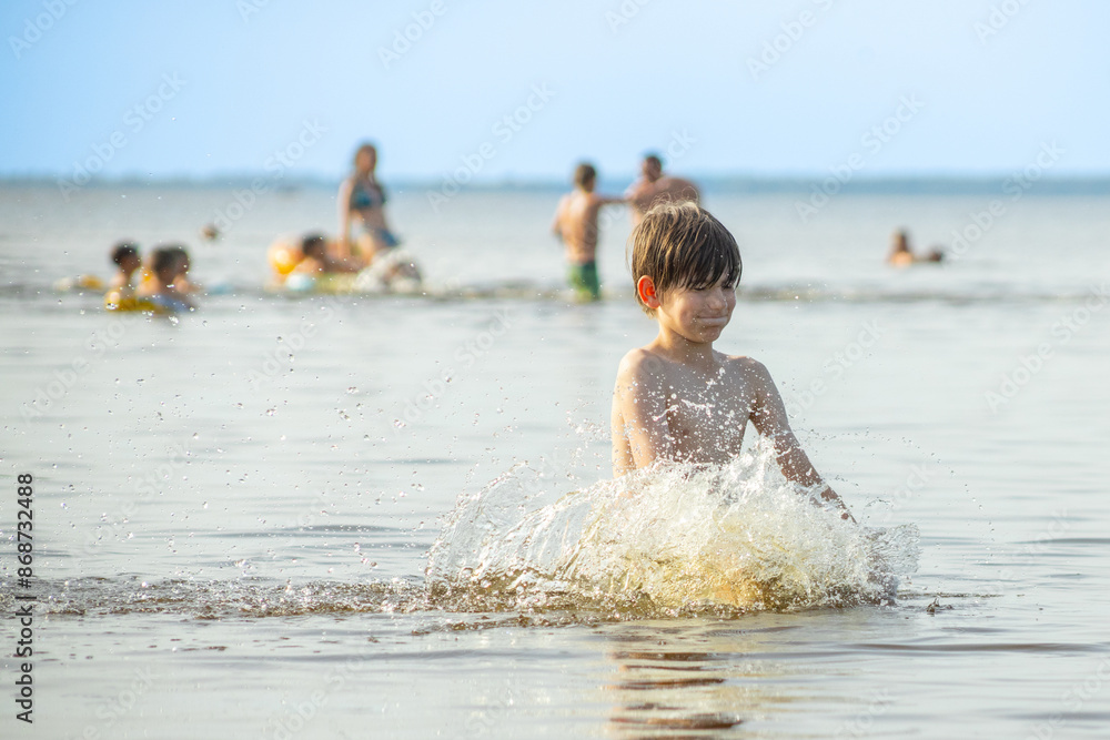 A young boy joyfully splashes water with both arms raised at the beach, creating a burst of droplets around him. Several people are visible in the background, enjoying the sunny day. A young boy joyfully splashes water with both arms raised at the beach, creating a burst of droplets around him. Several people are visible in the background, enjoying the sunny day.