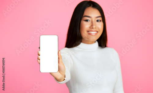 Asian young woman with long black hair wearing a white turtleneck sweater. She is holding a smartphone with a blank screen in front of her, looking directly at the camera with a bright smile