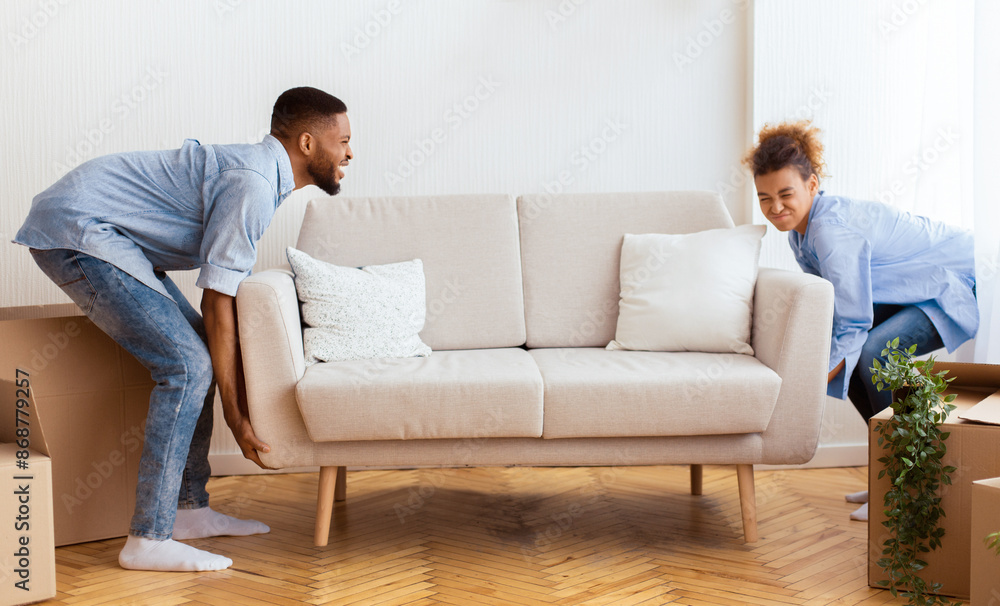 Relocation. Afro Couple Lifting Heavy Sofa Standing In Empty Room After Moving In New House Relocation. Afro Couple Lifting Heavy Sofa Standing In Empty Room After Moving In New House