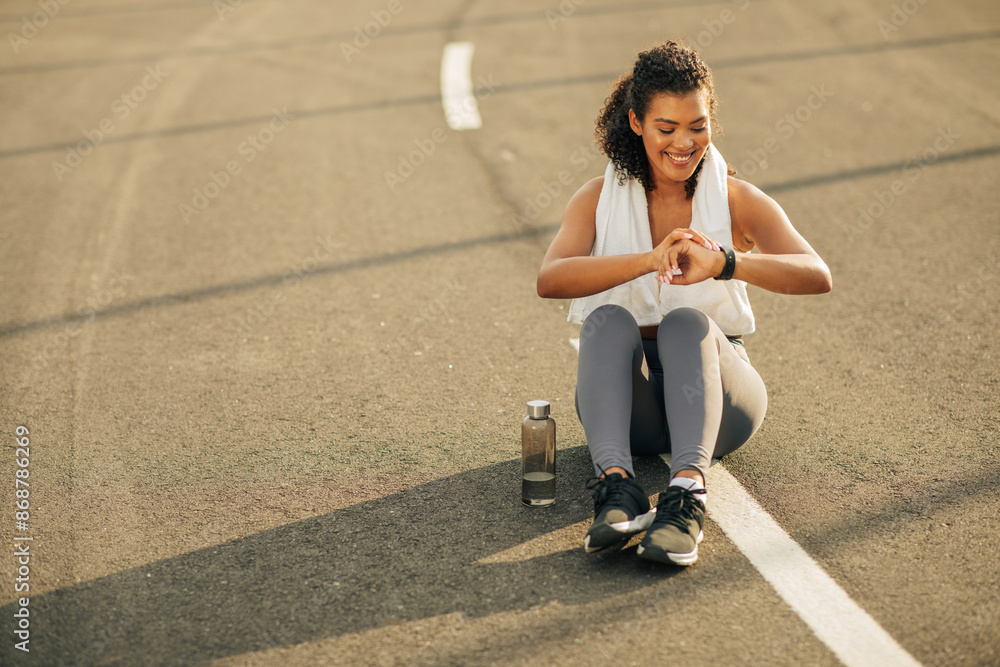 Fototapeta premium A young woman in athletic wear is sitting on a track, looking down at her fitness tracker. She is wearing a white towel around her neck and has a water bottle beside her