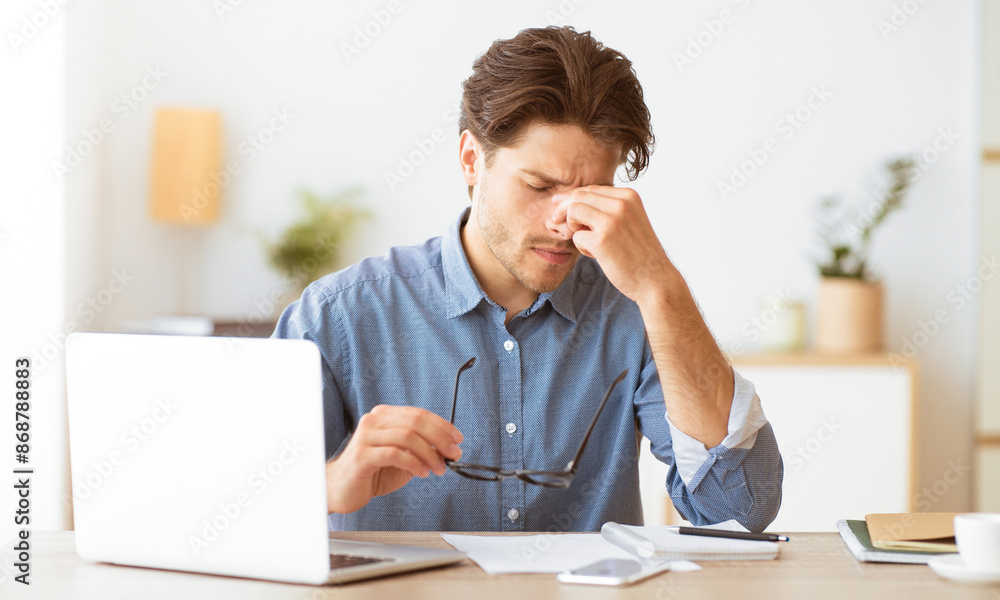 A man sits at a desk, rubbing his eyes in exhaustion. He is working on a laptop and appears to be experiencing eye strain. A man sits at a desk, rubbing his eyes in exhaustion. He is working on a laptop and appears to be experiencing eye strain.