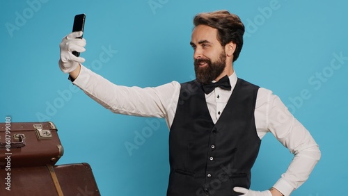 In studio, doorman in uniform takes photos while using smartphone for selfies and acting silly against blue background. Skilled hotel concierge does pictures with his gadget while on the job.