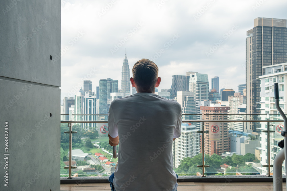 Fototapeta premium Rear view of man exercising on spinning bicycle on modern outdoor balcony overlooking Kuala Lumpur cityscape. Male doing cardio training on stationary bike on terrace of residental building