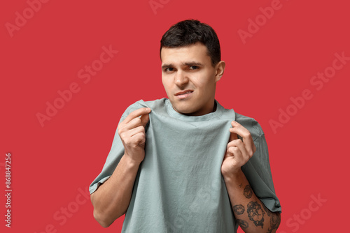 Handsome young man feeling bad smell from dirty clothes on red background photo