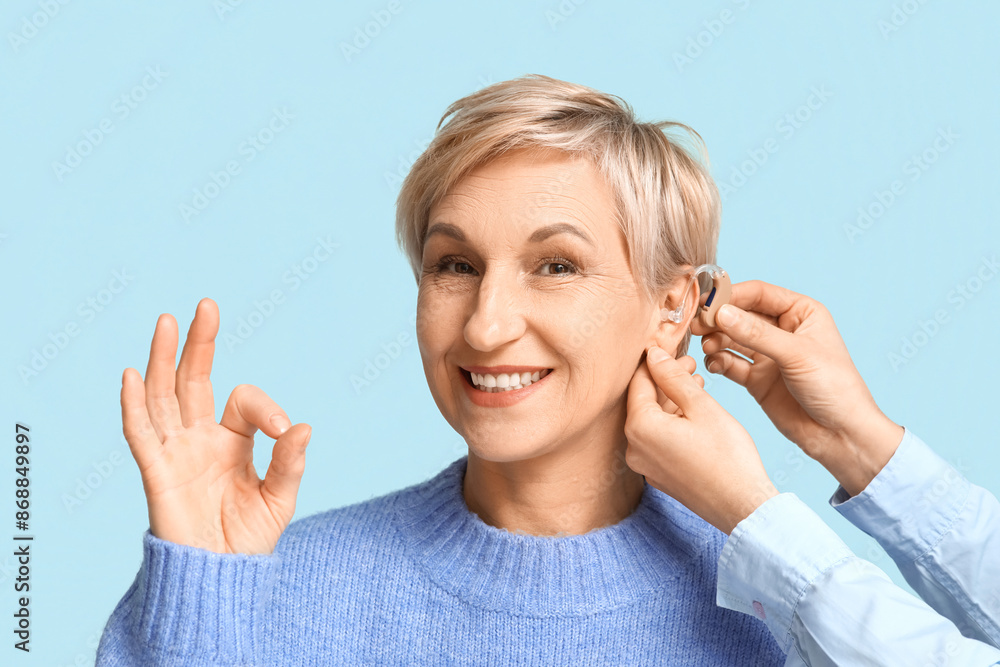 Obraz premium Young woman putting hearing aid in mature woman's ear showing ok gesture on blue background