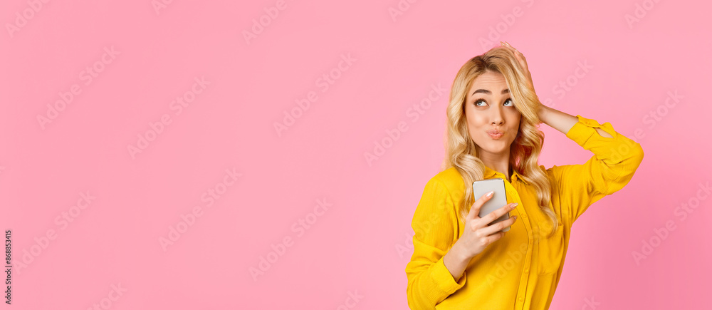 A woman wearing a yellow shirt is holding a smartphone in one hand while using the other hand to adjust her hair. She is standing in front of a pink background and looking at copy space A woman wearing a yellow shirt is holding a smartphone in one hand while using the other hand to adjust her hair. She is standing in front of a pink background and looking at copy space