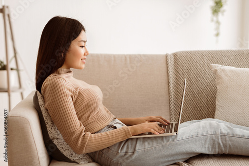A young woman sits on a beige couch working on a laptop computer.